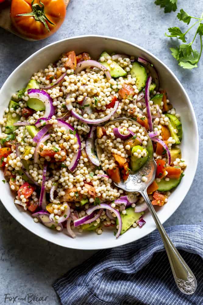 Israeli couscous salad in a white bowl with a serving spoon