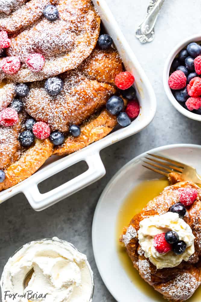Croissant French Toast Casserole in pan with berries and next to a single serving of croissant french toast on a plate.