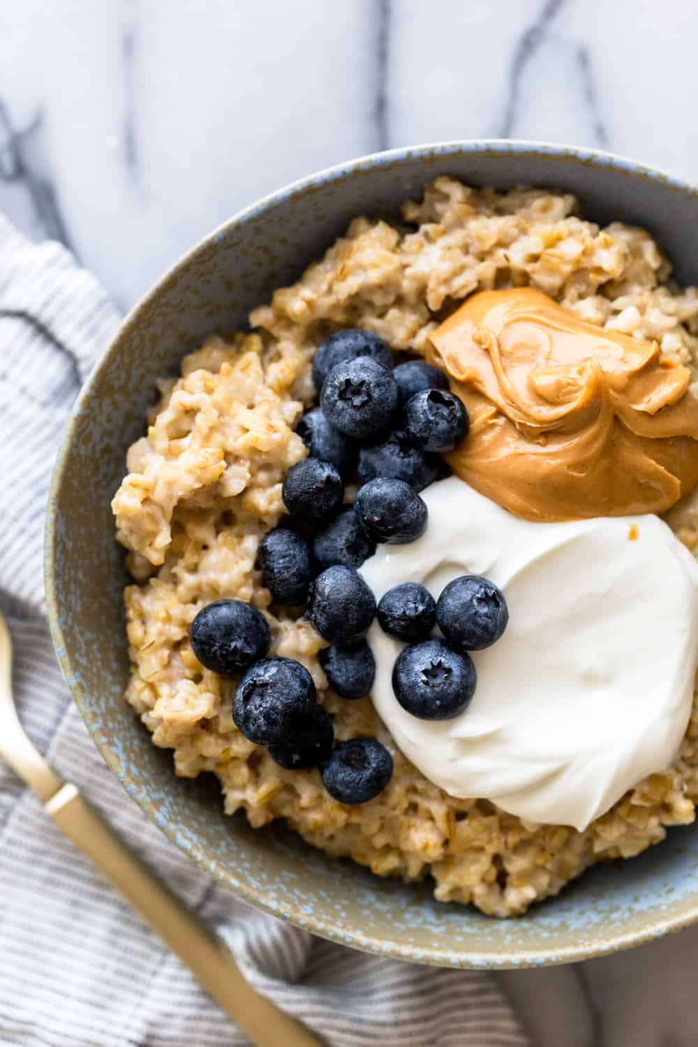 Oatmeal topped with yogurt, peanut butter and blueberries in a light blue bowl with a gold spoon