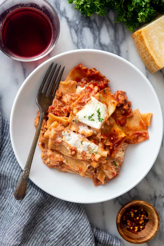 Lasagna in a white bowl with a marble background, next to a glass of wine