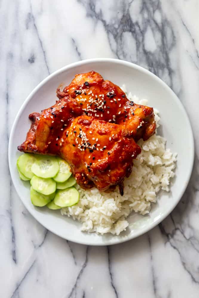 Korean chicken thighs on a white plate with rice and cucumbers