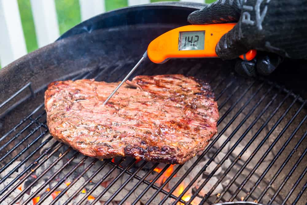 Flank Steak on a charcoal grill with a probe thermometer showing 142 degrees F