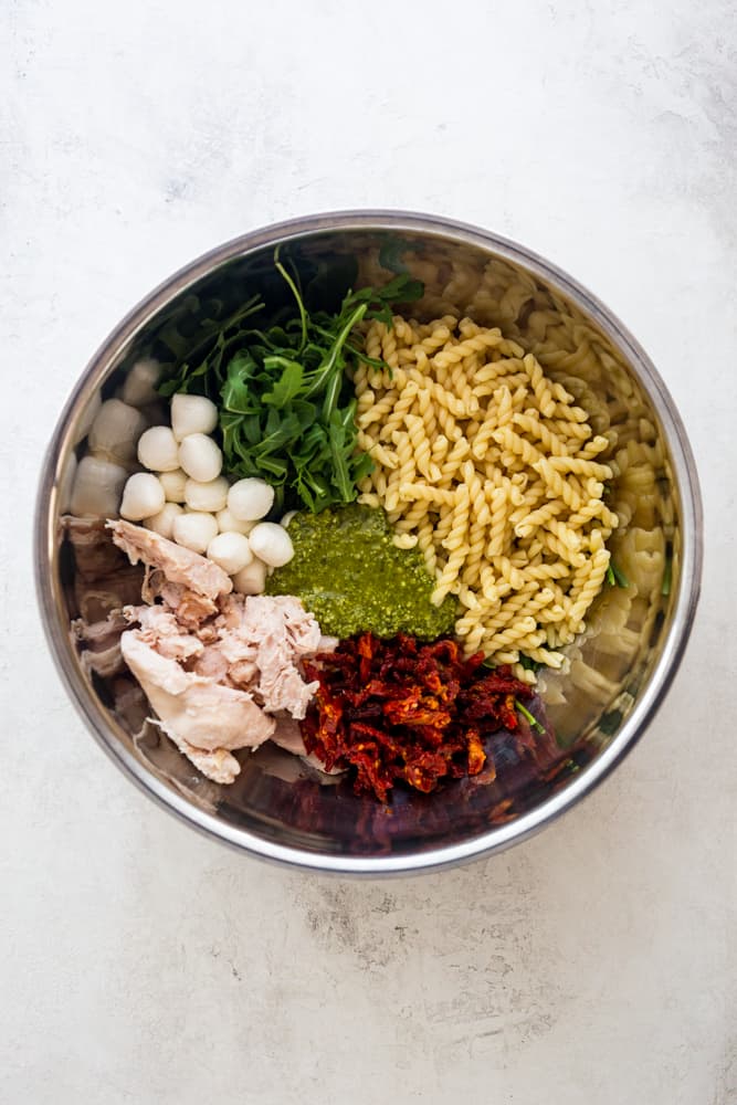 Ingredients for chicken pesto pasta salad - cooked pasta, arugula, mozzarella balls, cooked chicken, sun dried tomatoes, pesto - together in a large stainless steel bowl