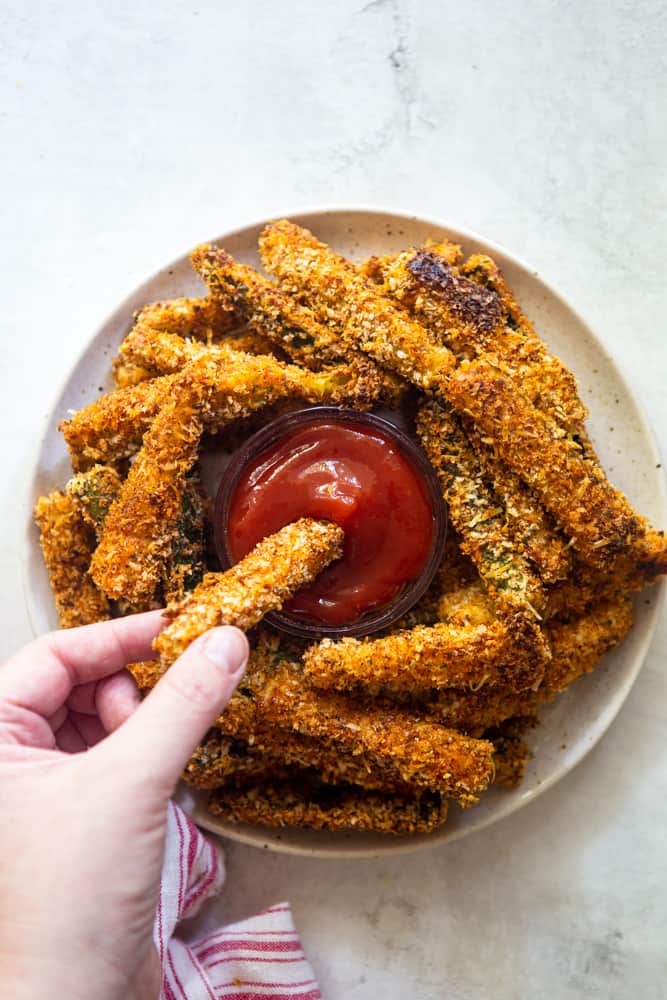 Plate of zucchini fries, a hand dipping on fry into ketchup