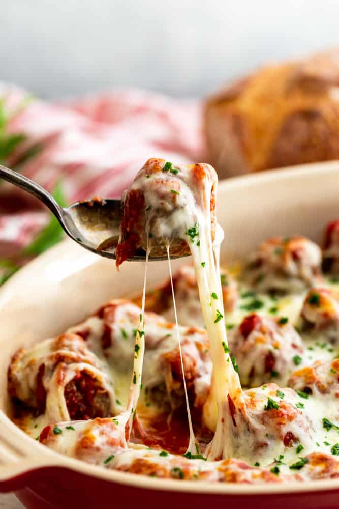 One meatball being lifted out of a pan of cheesy meatballs - with a gooey cheese pull.