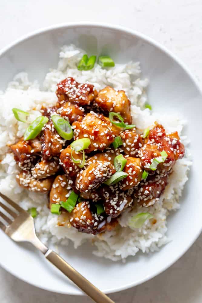Sesame chicken garnished with sesame seeds and sliced green onions over white rice, in a white bowl.