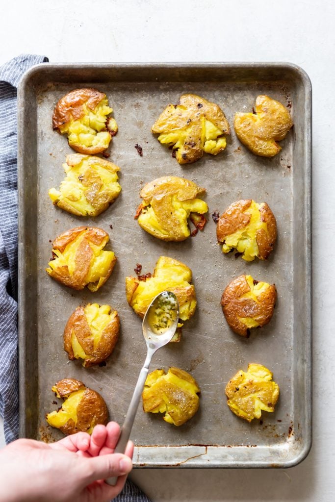 Smashed potatoes after baking, with a spoon of garlic herb butter about to be drizzles on top.