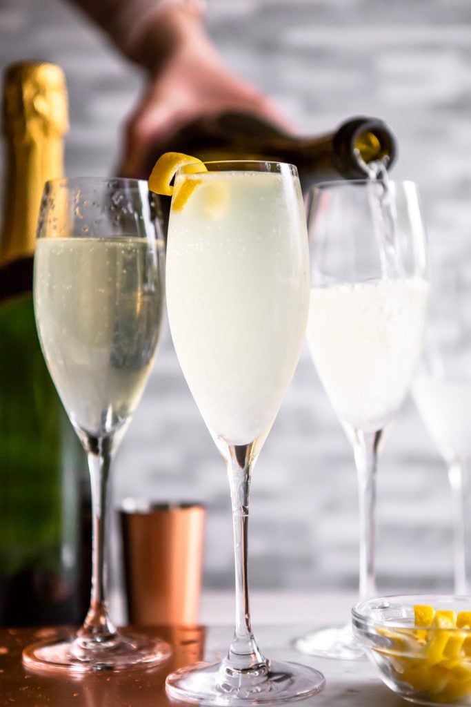 3 French 75 cocktails in champagne flutes, with a bottle of champagne being poured into one. 