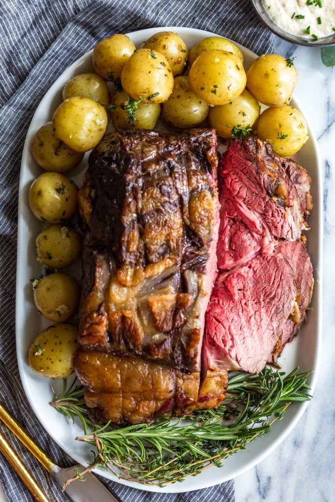 Slow Roasted prime Rib sliced showing pink interior, on a plate with baby potatoes and fresh herbs.