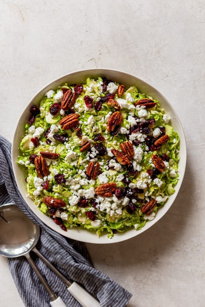 Shaved brussels sprouts salad with goat cheese, dried cranberries and candied pecans, in a while bowl next to a blue dishtowel.