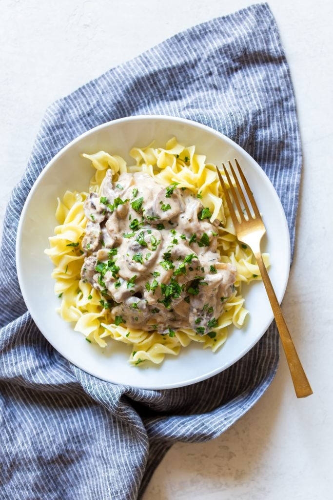 Creamy beef stroganoff over egg noodles' in a white bowl, on a blue striped dish towel. 