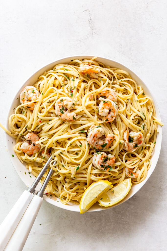 Shrimp scampi pasta in a white bowl with white serving utensils.