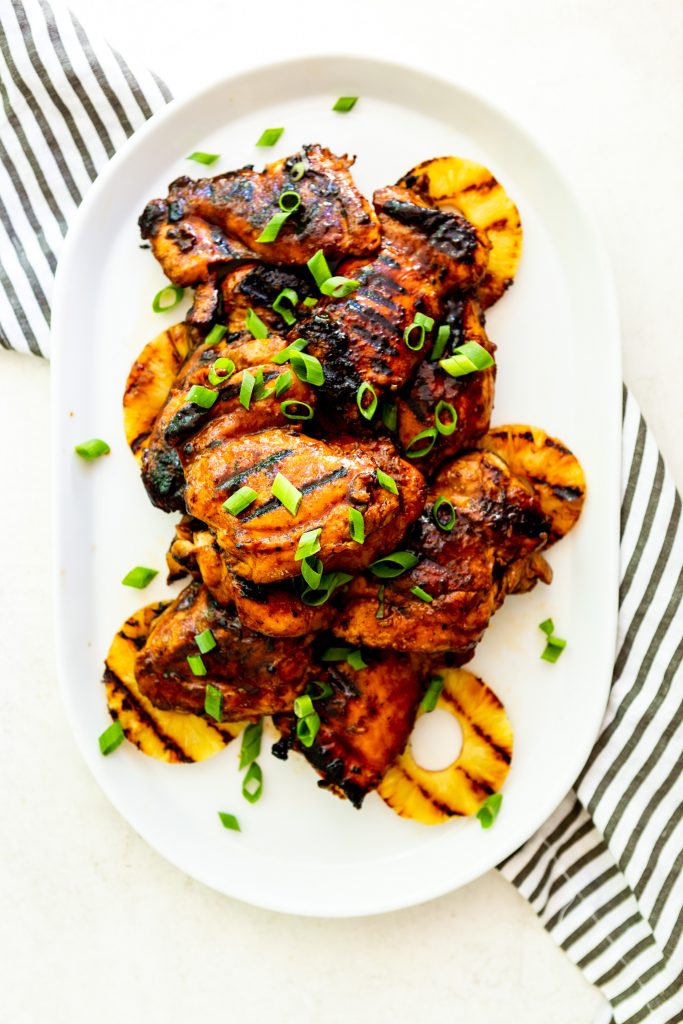 Grilled huli huli chicken thighs and grilled pineapple piled on a white platter, garnished with green onions.