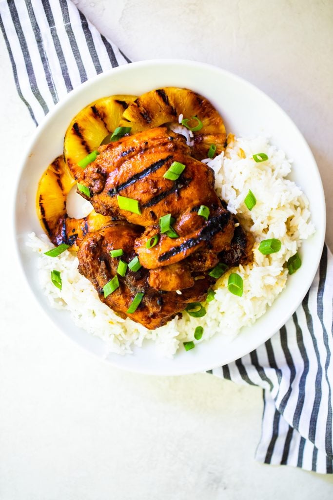 A plate of huli huli chicken on top of rice and grilled pineapple, garnished with green onions.