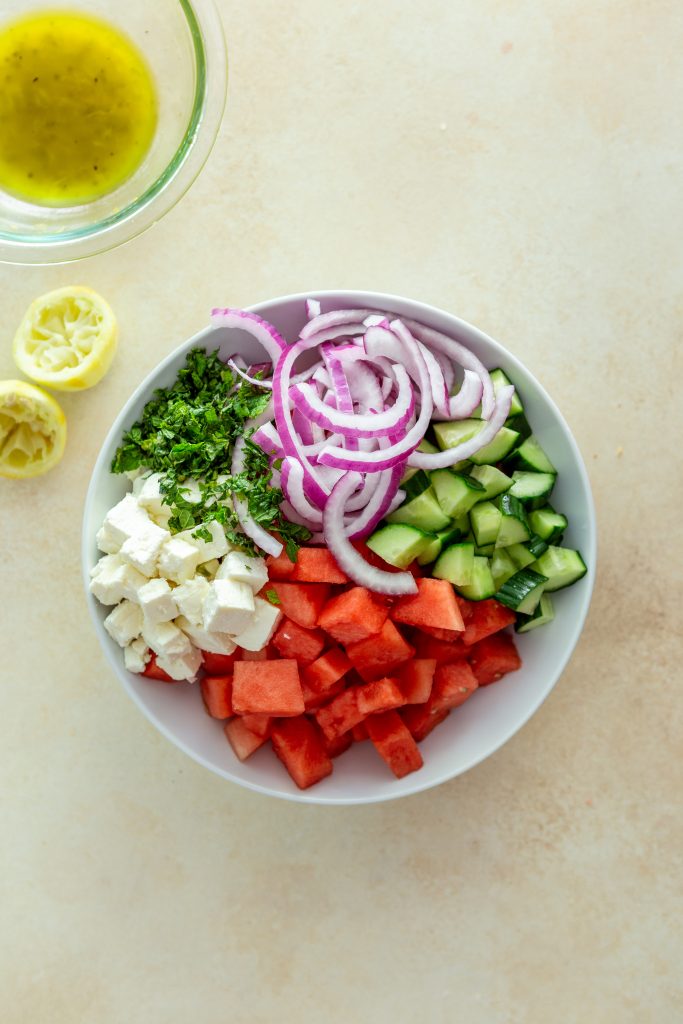 Chopped mint, Sliced red onions, chopped cucumber, cubed watermelon, cubed feta, all in a bowl before being mixed.