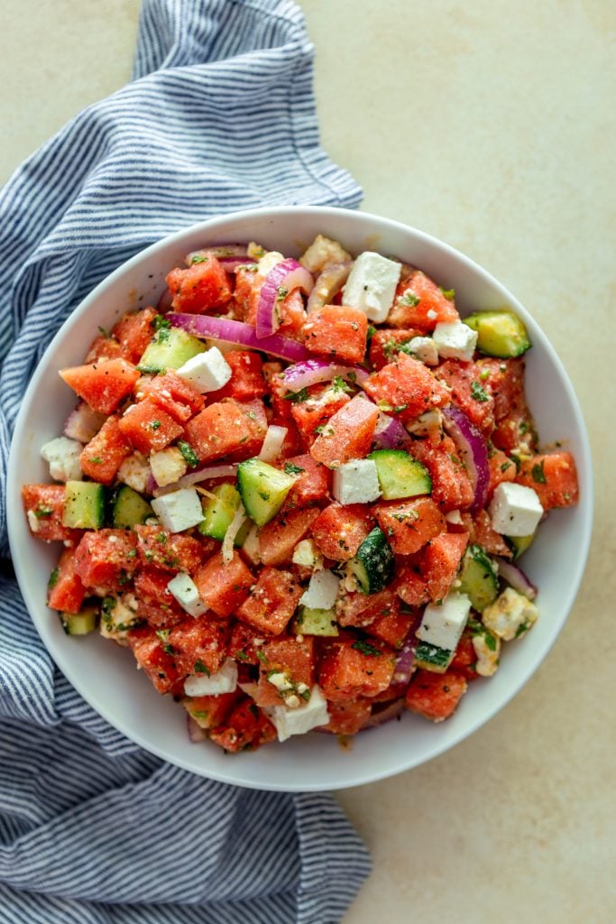 Watermelon Feta Salad with red onions, cucumbers and mint in a white bowl with a blue and white striped kitchen towel nearby.