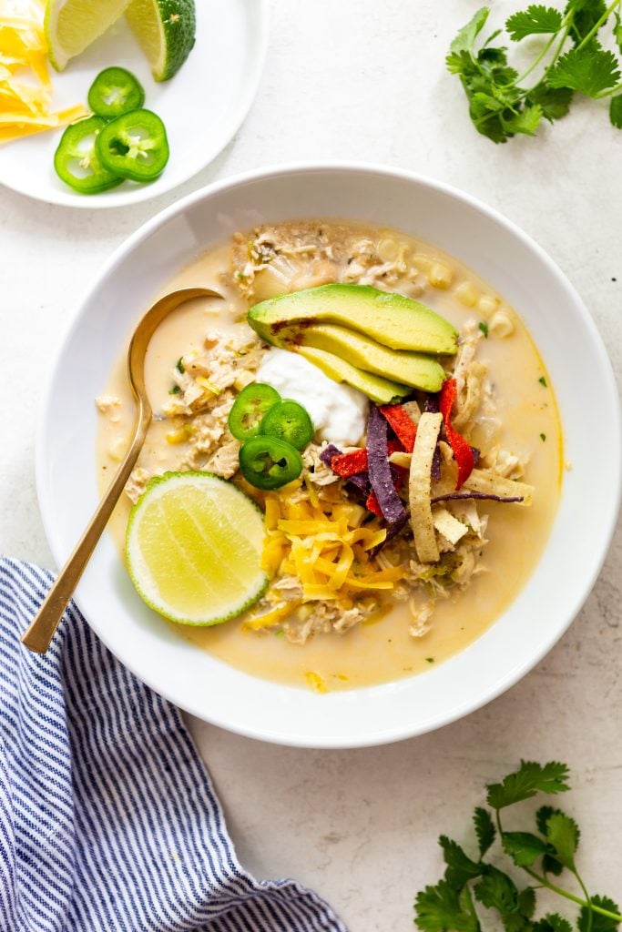 Creamy white chicken chili in a white bowl, topped with avocado slices, sour cream, shredded cheese, tortilla strips, jalapeno slices and a lime wedge.