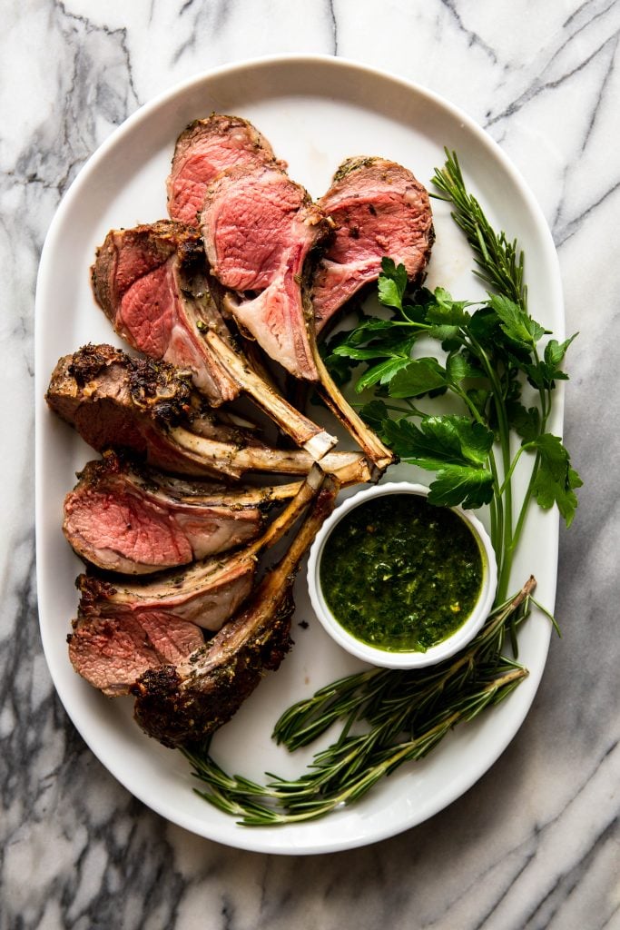Roasted lamb chops on a white platter with herbs and chimichurri sauce.