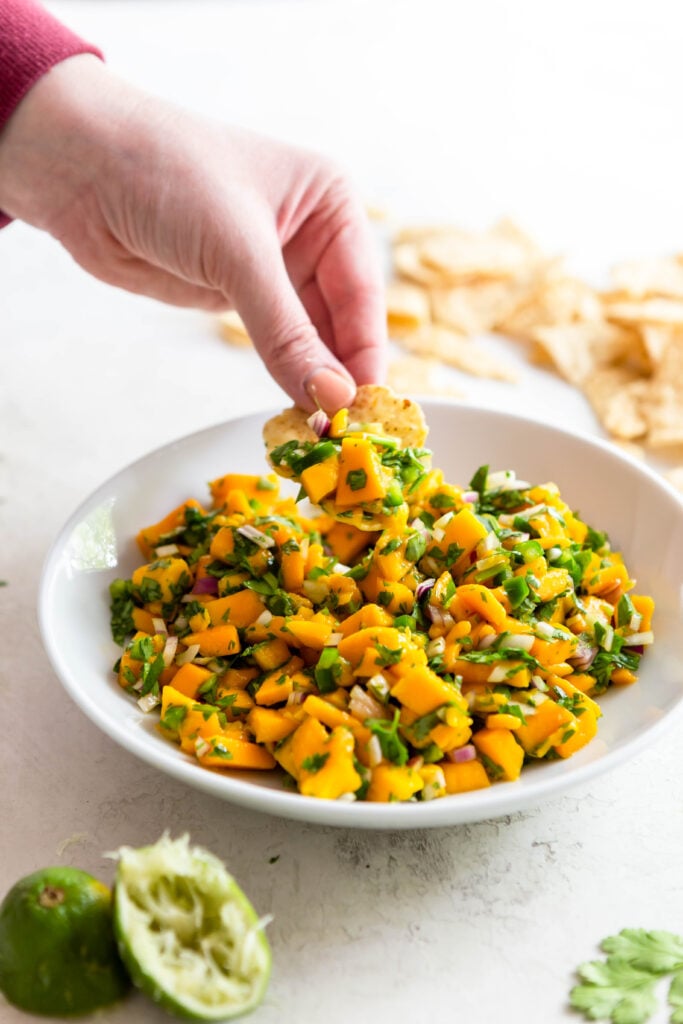 Hand holding a corn chip scooping mango salsa out of a white bowl. 