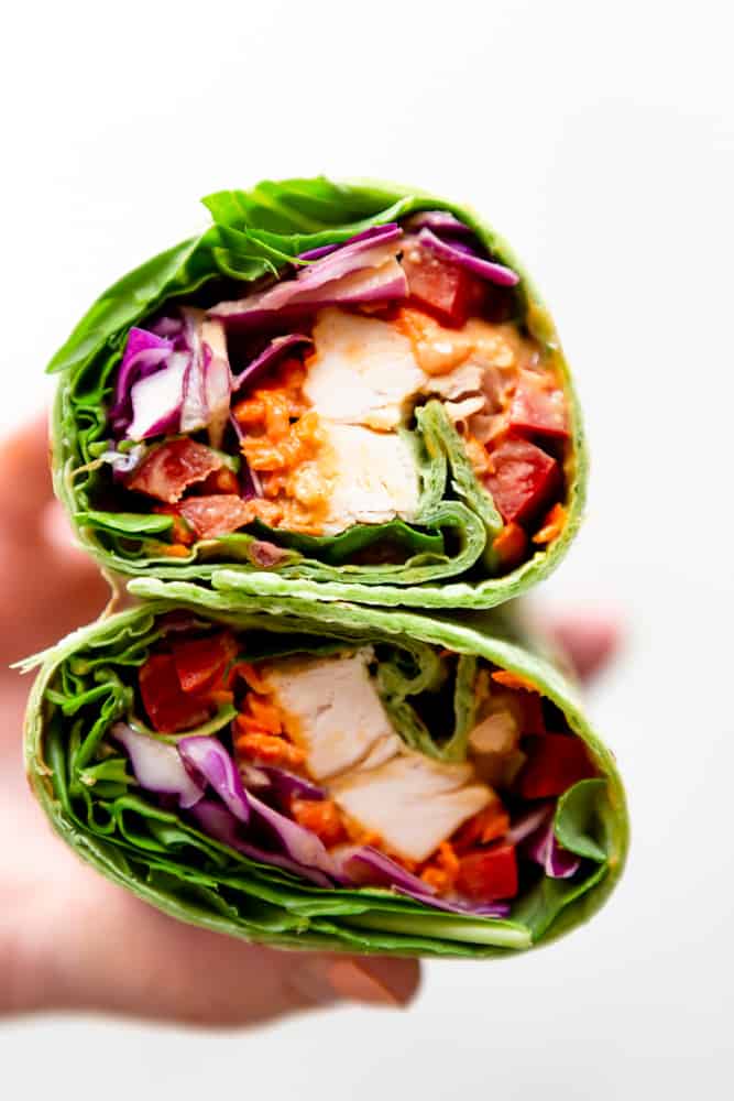 Close up of a peanut chicken wrap cut in half, showing chicken, bell pepper, cabbage, carrots, spinach and peanut sauce in a green spinach wrap. 