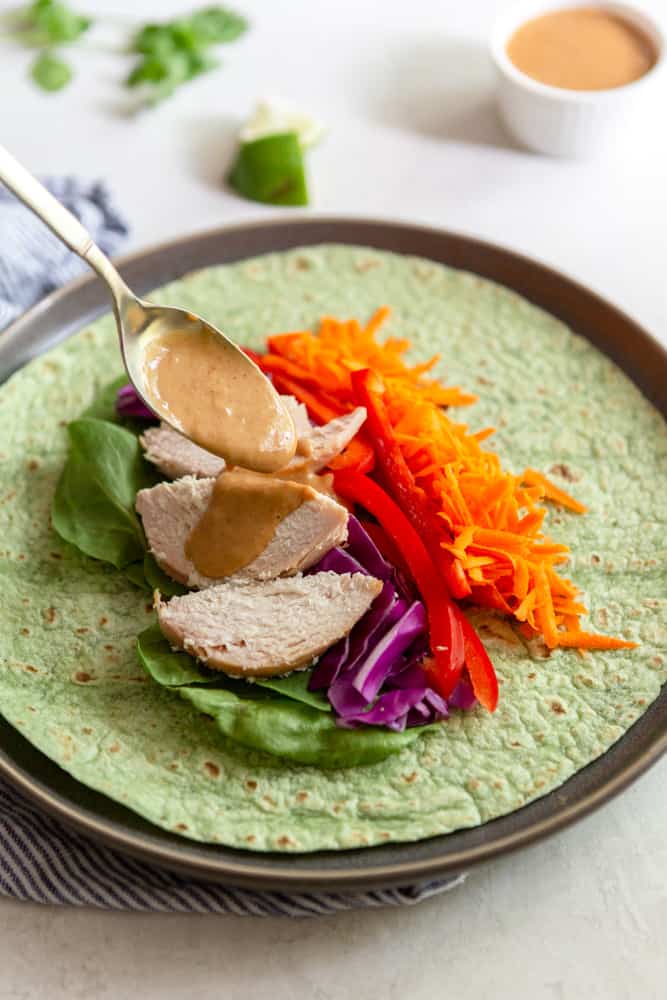 Spinach tortilla with spinach, cabbage, red bell pepper and carrots, with a spoonful of peanut sauce being drizzled on top.