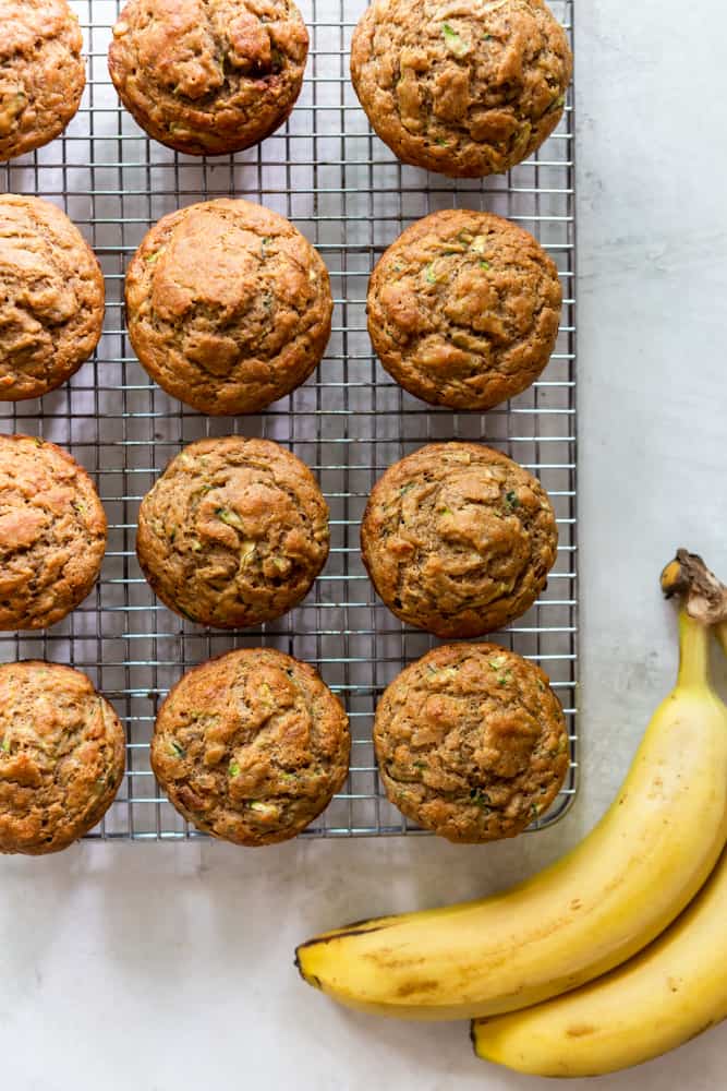Overhead shot of banana zucchini muffins on a wire rack. on a white background, next to some bananas.