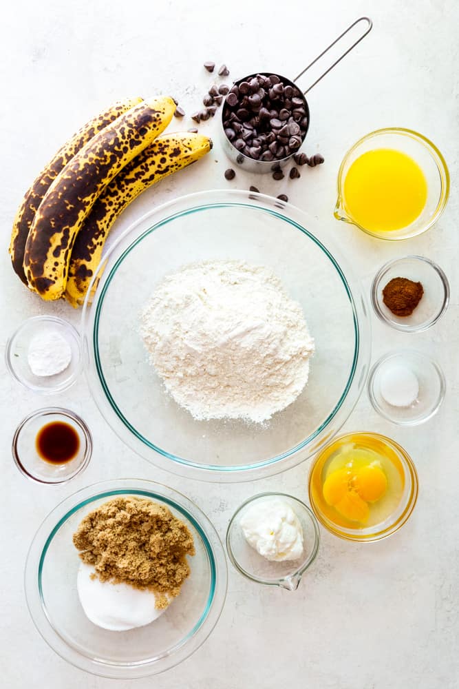 Ingredients for banana bread - a glass bowl of flour surrounded by three overripe bananas, chocolate chips, melted butter, cinnamon, salt, eggs, yogurt, brown and white sugar, vanilla and baking soda.