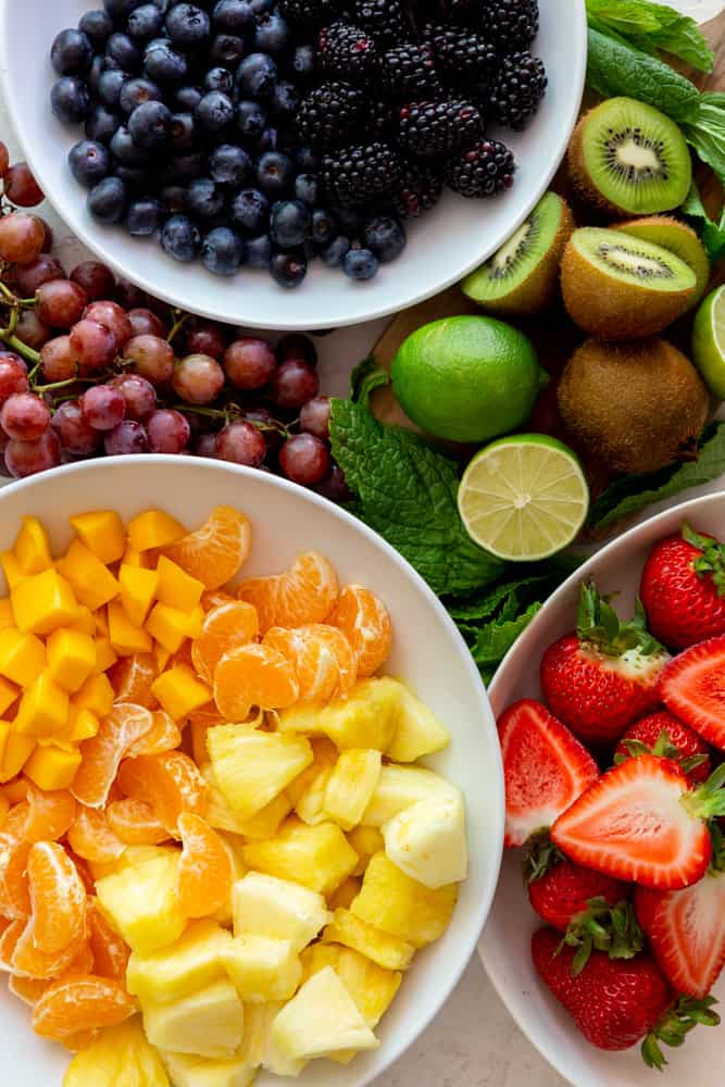 Ingredients for fruit salad blackberries and blueberries in a white bowl, grapes on a white surface nest to the blueberries limes, kiwi and mint on a cutting board, mango, tangerine and pineapple in a white bowl, strawberries in a white bowl.