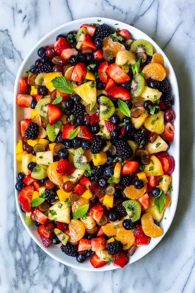 A beautiful rainbow fruit salad garnished with mint on a white platter, on a marble backdrop.