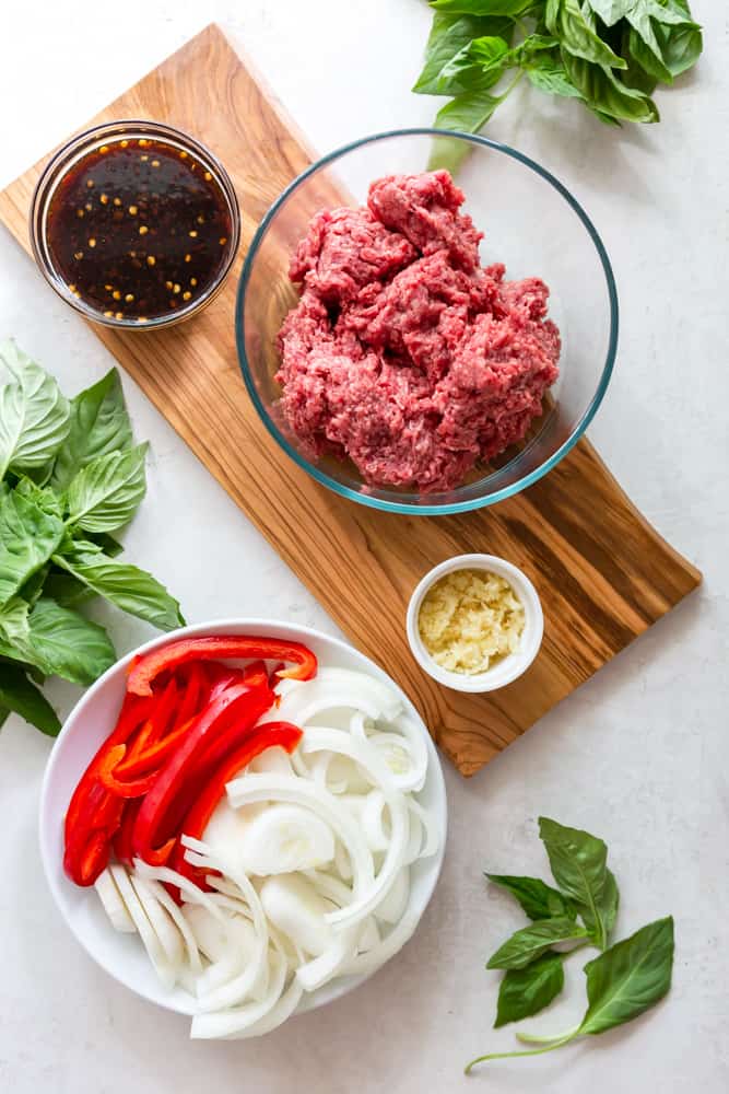 Ingredients for Thai basil beef - a bowl with the sauce, ground beef, minced garlic, red bell peppers and onions, surrounded by basil.