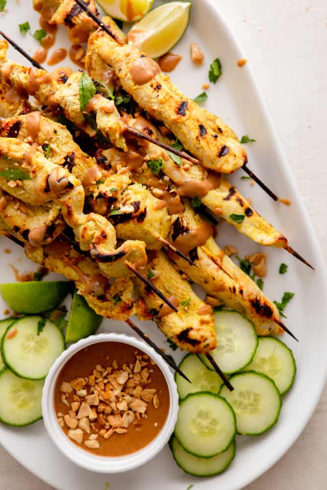 Close up of chicken satay skewers, garnished with peanut sauce, peanuts, cilantro, and lime.  A small bowl of peanut sauce and sliced cucumbers are on the side.