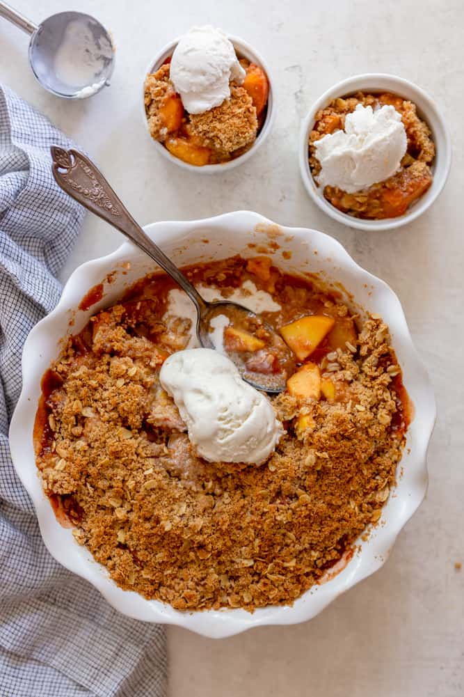 Peach crisp in white round pan, topped with ice cream.  Two small dishes of each crisp are next to it. 