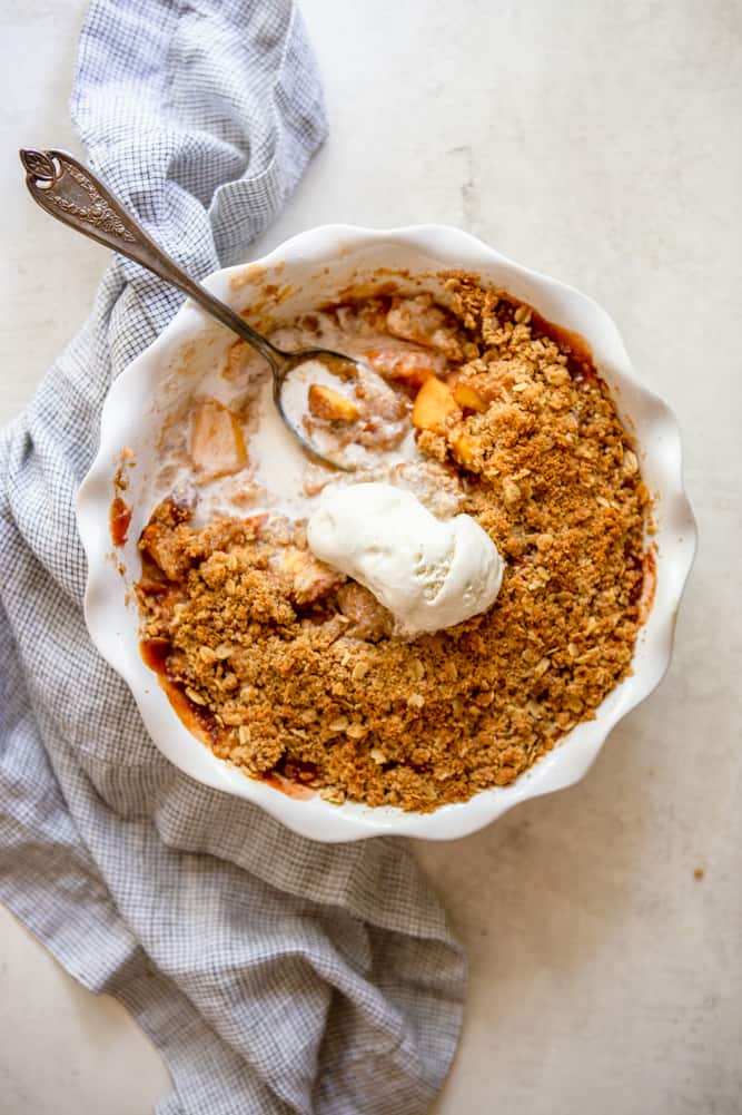Partly eaten peach crisp, topped with vanilla ice cream that is starting to melt.