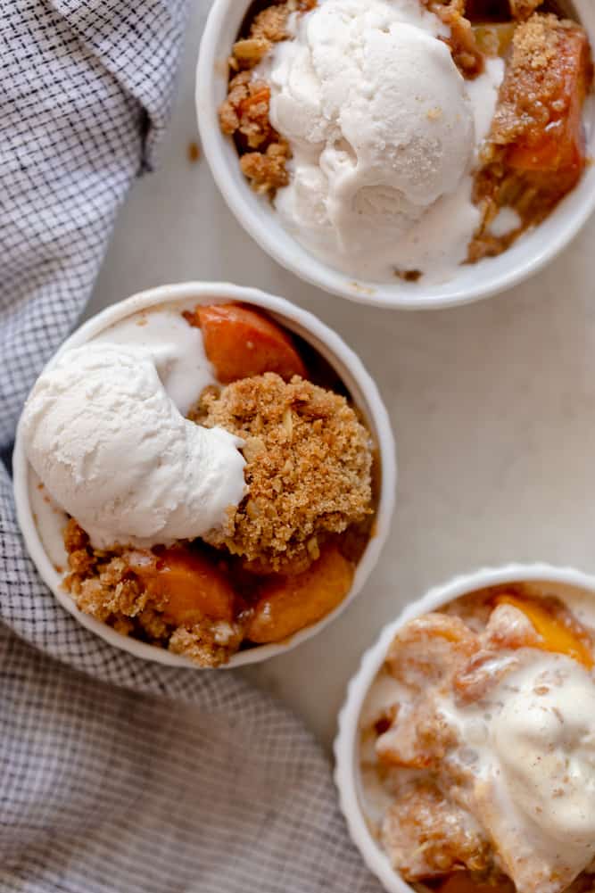 Individual peach crisp servings in white ramekins, topped with vanilla ice cream.