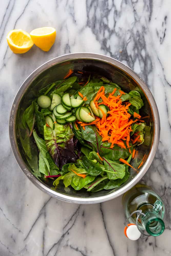 Mixed greens with shredded carrots and sliced cucumbers in a metal mixing bowl.