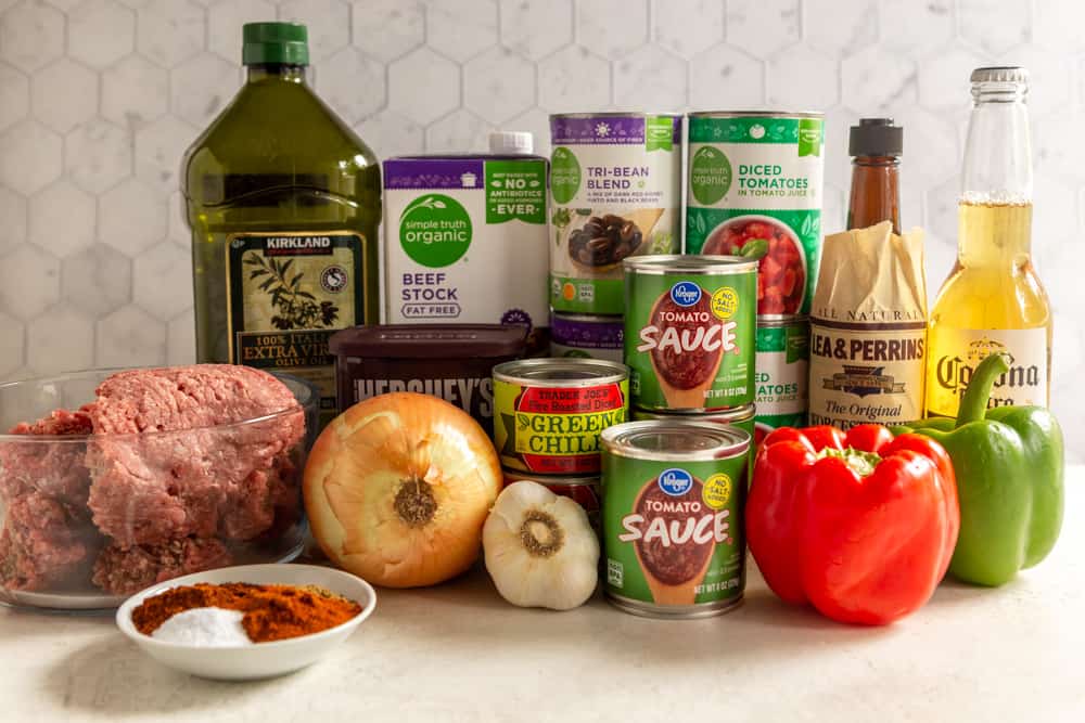 Ingredients for chili - Ground beef, olive oil, beef stock, canned beans, canned diced tomatoes, Worcestershire sauce, beer, onion, garlic, canned green chilis, canned tomato sauce, red bell pepper, green bell pepper, a small white dish with spices and salt.