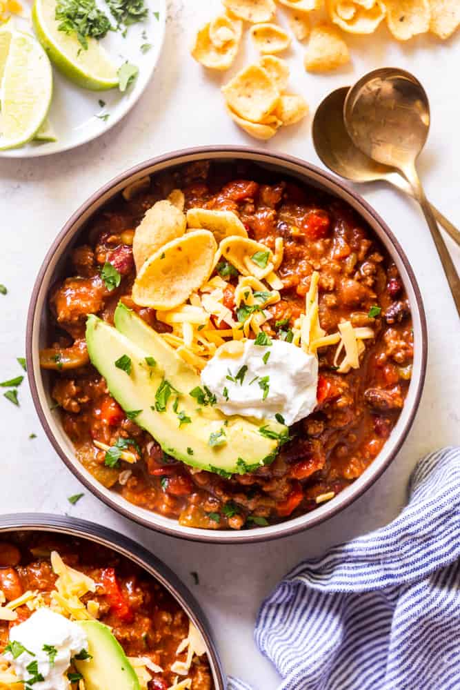 A bowl of slow cooker chili topped with avocado, corn chips, grated cheese and sour cream, garnished with parsley.