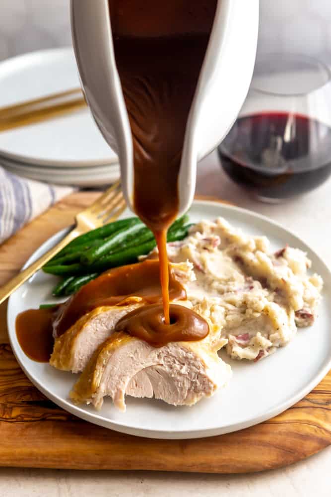 White gravy boat pouring gravy onto sliced turkey with mashed potatoes and green beans.