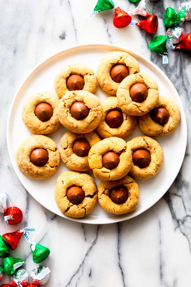 Peanut butter blossom cookies on a white plate, surrounded by red, green and silver wrapped Hershey kisses.