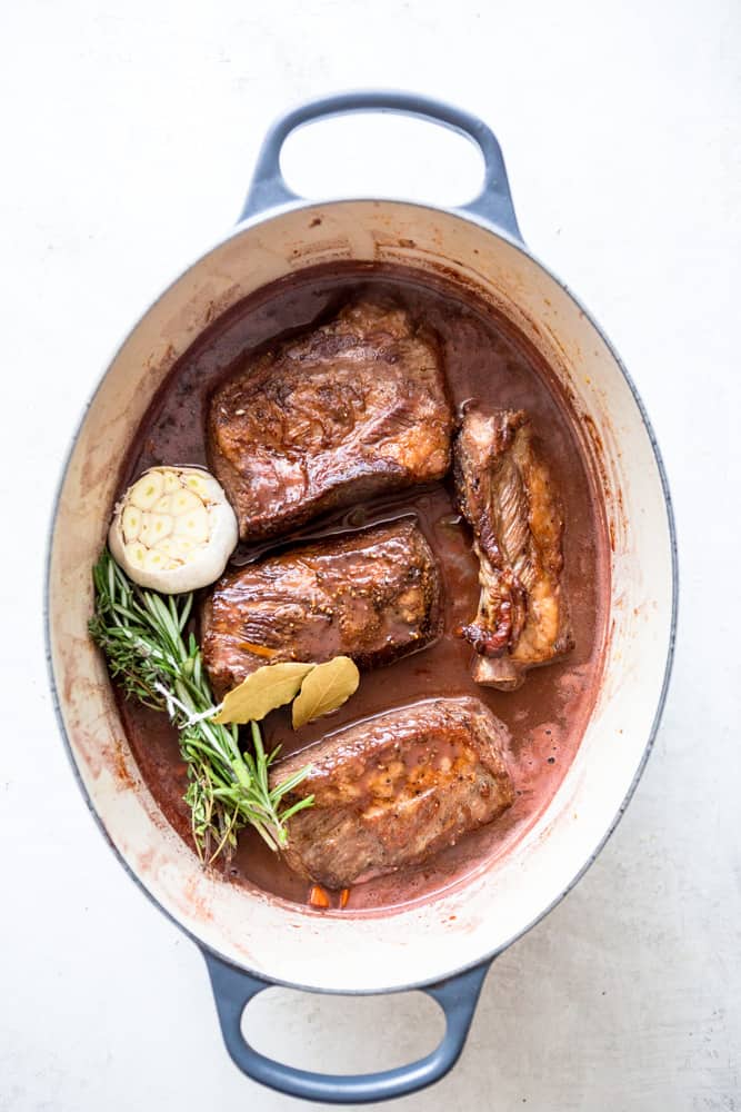 A large, oval shaped dutch oven with short ribs that have been browned, a bundle of herbs, a head of garlic cut in half, two bay leaves, and red wine braising liquid - before cooking.