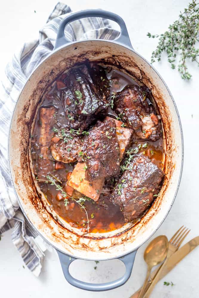 A large, oval shaped Dutch oven with red wine braised short ribs garnished with thyme