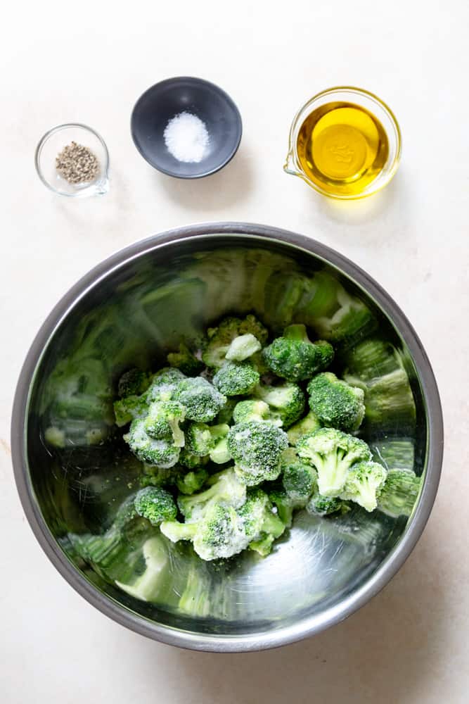 Frozen broccoli in a stainless steel bowl, small dishes of olive oil, salt and pepper nearby.