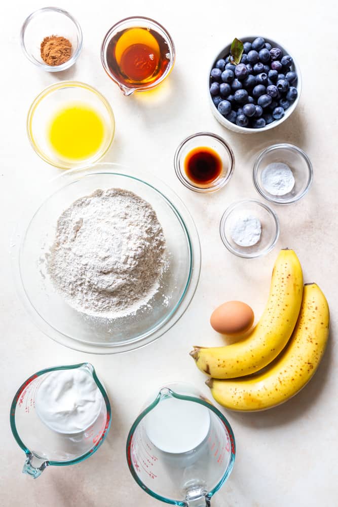 Ingredients for healthy blueberry banana muffins: White whole wheat flour, baking soda, baking powder, cinnamon, vanilla extract, milk, yogurt, 2 ripe bananas, melted butter, maple syrup, fresh blueberries.