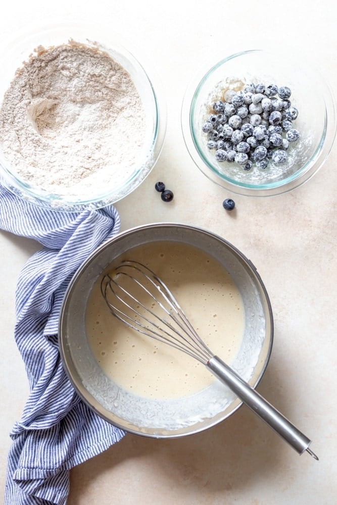 A bowl of dry ingredients, a bowl of wet ingredients and a bowl of blueberries that have been tossed in flour.
