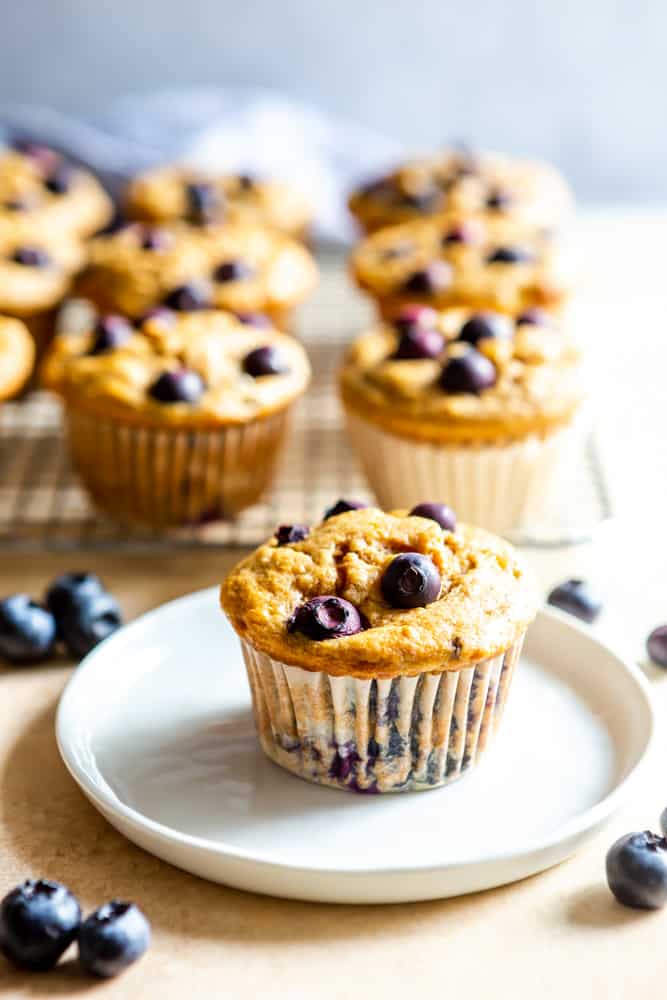 A healthy blueberry banana muffin on a white plate, with more muffins on a cooling rack behind it.