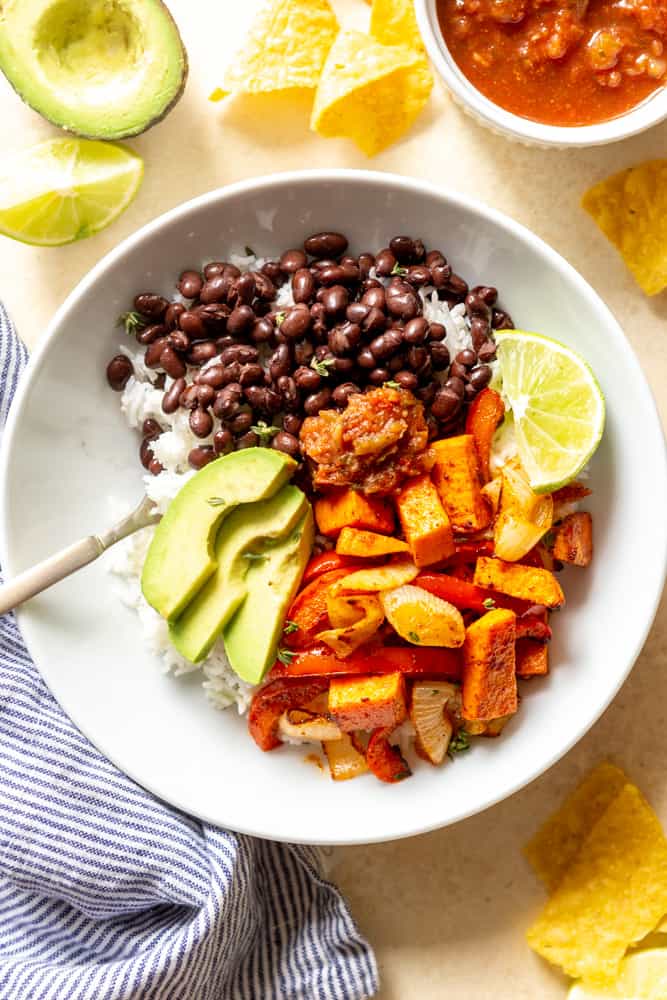 Veggie burrito bowl with white rice, black beans, roasted sweet potatoes, bell pepper and onions, with avocado and salsa in a white bowl. 