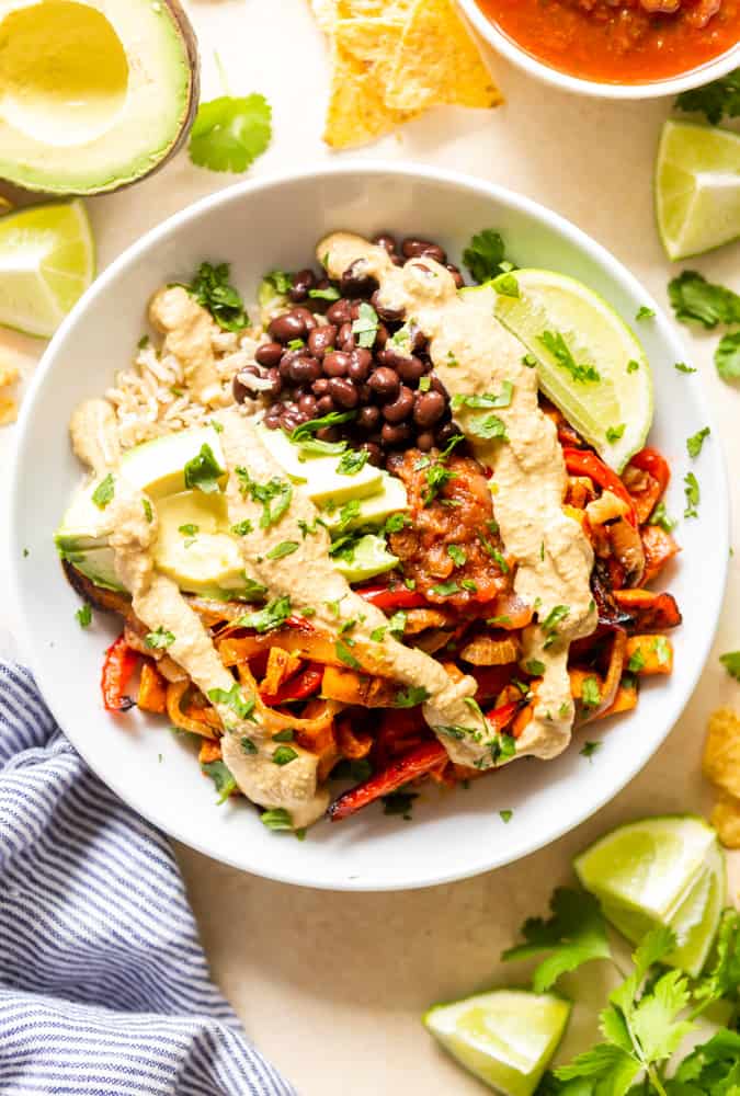 Roasted veggie burrito bowl with brown rice, roasted sweet potatoes, onions and bell peppers, black beans, sliced avocado, salsa and vegan queso in a white bowl.