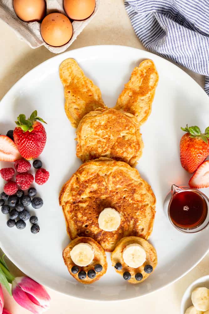 Pancakes in the shape of a rabbit with a slice of banana for a tail and banana slices for feet with blueberries for toes.