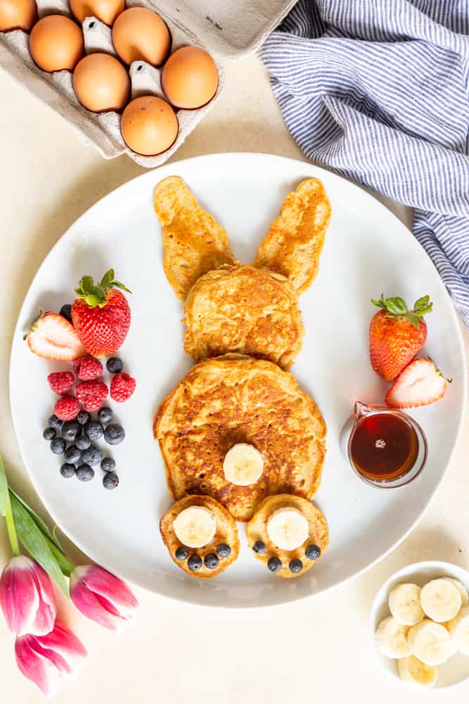 Carrot cake pancakes in the shape of a bunny - a large pancake for a body, a smaller pancake for a head, two triangular pancakes as ears. two mini pancakes as feet with banana slices and blueberries as the paw, and a banana slice as the tail.