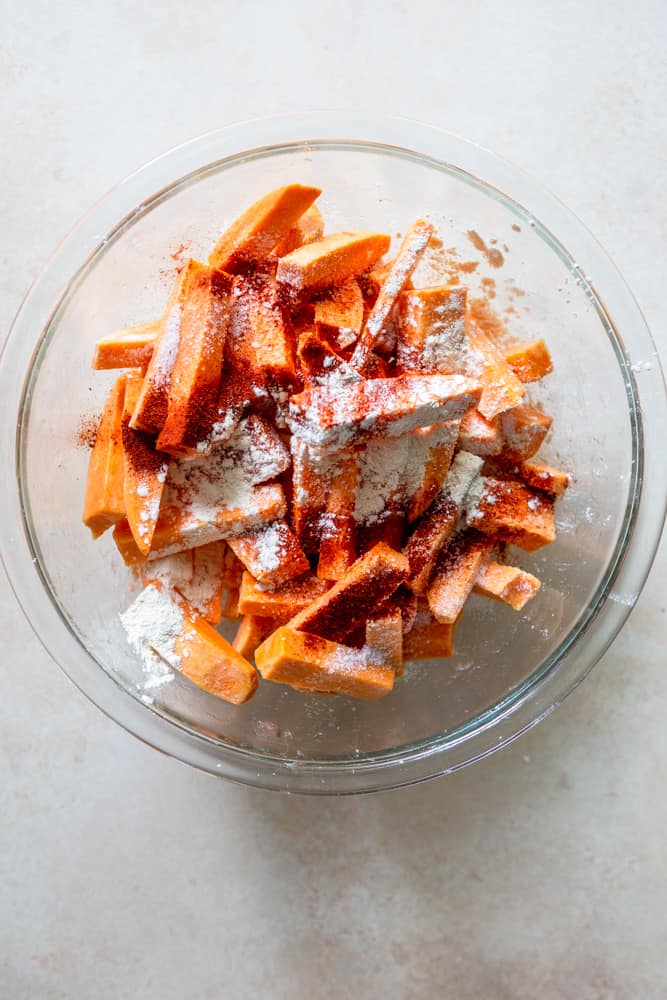 uncooked sweet potato sticks in a glass bowl with cornstarch, paprika and spices.